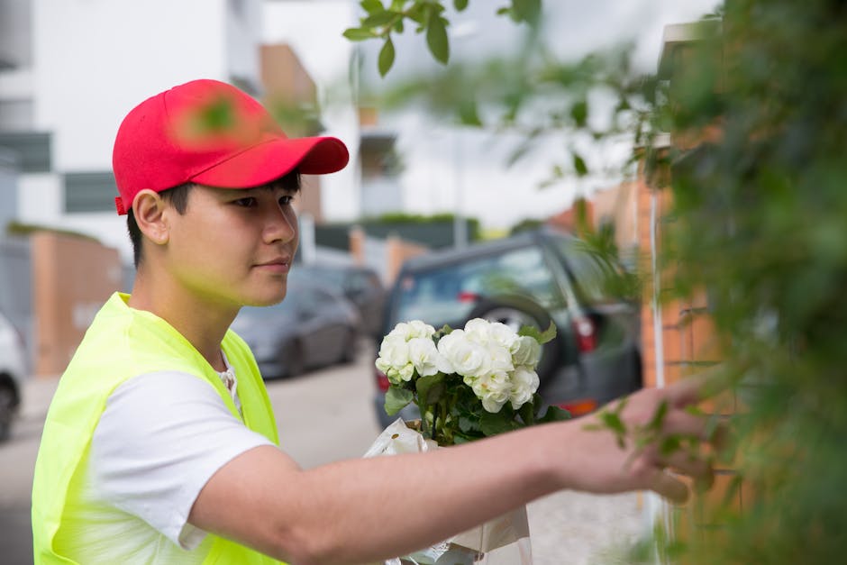 A young male florist wearing a red cap and bright yellow safety vest is seen arranging a bouquet of fresh white hydrangeas wrapped in light brown paper. He is outdoors, standing near a brick wall part