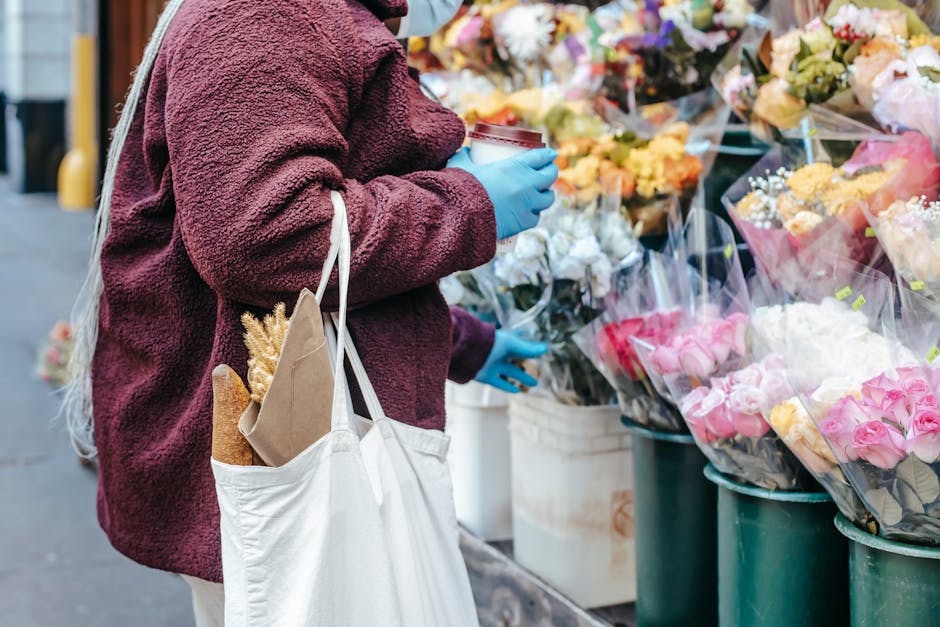 A person wearing a burgundy fleece jacket and blue disposable gloves is standing at an outdoor flower stall, holding a cup of coffee in one hand while selecting flowers with the other. They carry a wh
