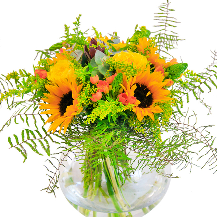 The initial impact of this arrangement is a wave of luminous, sun-inspired brilliance, reminiscent of the golden hours in Barking's town centre. Glorious sunflowers-bold and expressive-occupy centre stage, their oversized yellow petals shining with positivity beside the tightly layered rosettes of buttery yellow garden roses. Each bloom seems handcrafted with the artisan touch of a Barking florist, layered to create a sense of depth and abundance. Around these central flowers, droplets of glossy, crimson hypericum berries sparkle, offering a bridge between the cheerful yellows and the lush deep green foliage. Fern fronds trail gently down the sides, echoing the gentle sway of foliage seen along the Abbey Road gardens, while airy filler and delicate eucalyptus leaves introduce subtle shifts of light green and silvery blue, enhancing the arrangement's modern, yet timeless, appeal. The clear, spherical glass vase acts as a showcase, revealing bundles of green stems and intertwining textures beneath the surface, the water glimmering as it catches the light. Subtle plays of shadow add even more life, hinting at the bouquet's sculptural design. Its fragrance lingers with notes of newly-cut blooms and outdoorsy freshness, blending beautifully into a Barking morning or evening. This arrangement exudes a spirit of happiness and celebration-perfect for marking birthdays, congratulating a friend, or bringing a special touch to a Barking home or office. Every thoughtful detail in the bouquet is a testament to skilled local florist craftsmanship and an understanding of what makes the Barking community unique.